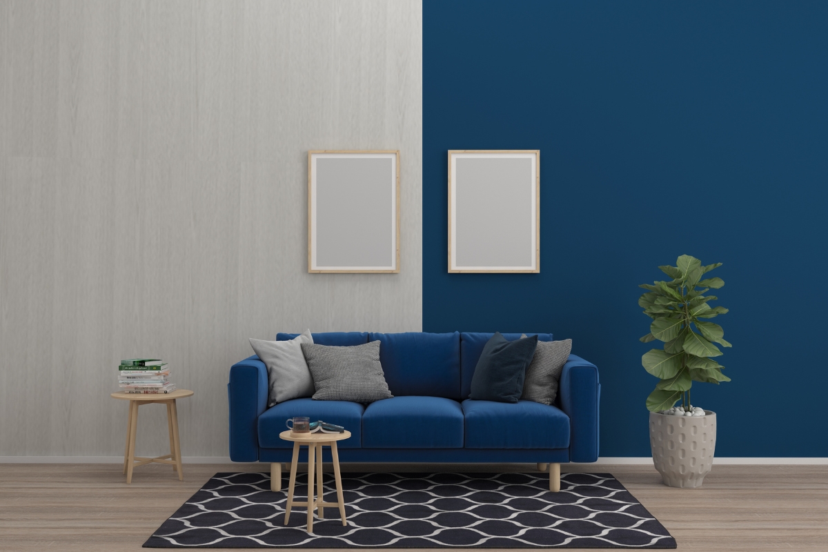 A modern living room showcases the latest home interior design trends with its blue sofa, assorted pillows, a small side table with books, a potted plant, a geometric rug, and two blank picture frames on a two-toned wall.
