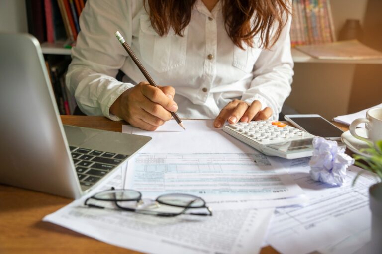A person in a white shirt is sitting at a desk, diligently budgeting for home improvements with a pencil and a calculator. A laptop, glasses, crumpled paper, and a cellphone are also on the desk.