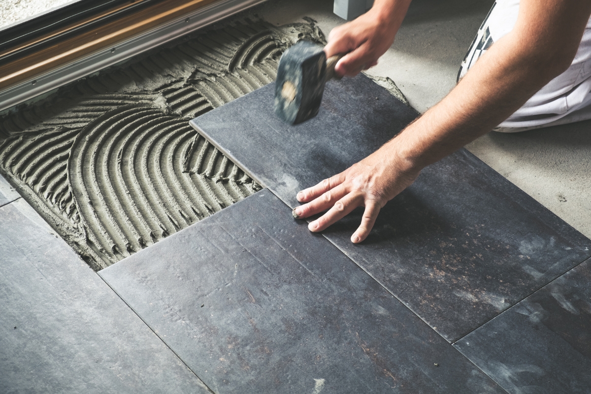 Budgeting for Home Improvements: Savvy Tips for Flooring Costs 4 A person is installing grey floor tiles with adhesive cement and using a rubber mallet, mindful of budgeting for home improvements.
