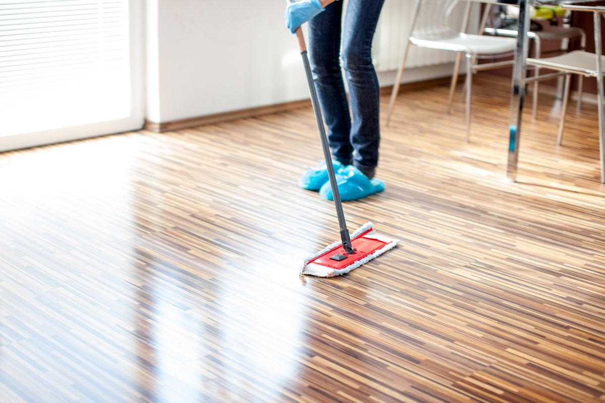 A person dedicated to floor maintenance is mopping a wooden floor in a bright room, wearing blue shoe covers and gloves.