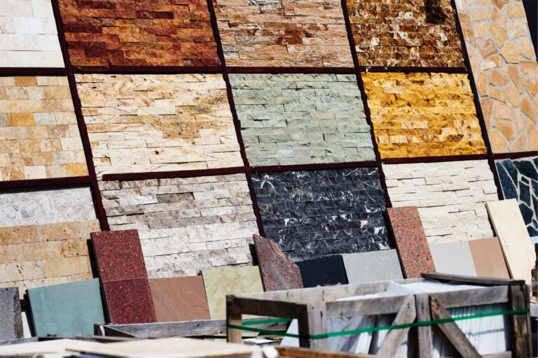 Various samples of stone and tile cladding, along with a selection of flooring materials, are displayed on a vertical board outdoors with some crates in the foreground.