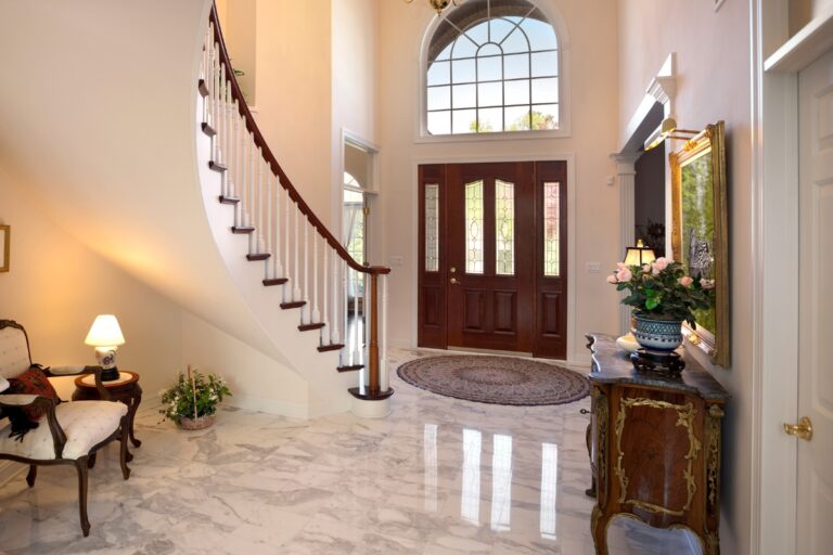 A bright foyer with marble luxury home flooring, a curved staircase, wooden double doors, a round rug, and ornate furniture accented by a floral arrangement.