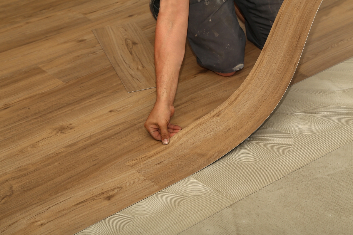 Luxury Home Flooring Ideas for Buckhead: Hardwood, Tile, and More 3 Person installing wood-look vinyl flooring, aligning a plank over adhesive on the floor. Only the arms and legs of the installer are visible, highlighting the careful installation of luxury home flooring.