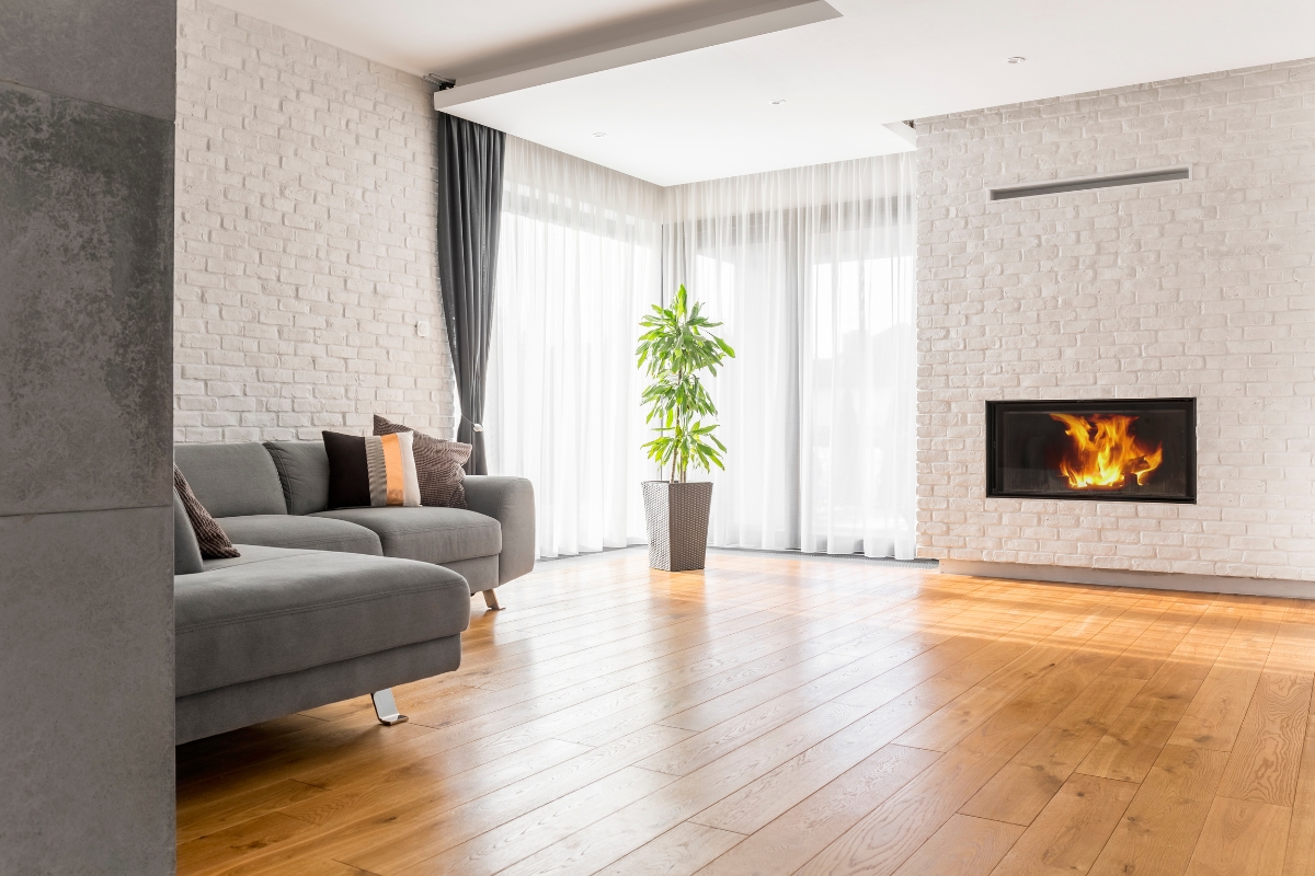 Luxury Home Flooring Ideas for Buckhead: Hardwood, Tile, and More 6 Modern living room with luxury home flooring, grey sectional sofa, potted plant, white brick walls, large windows with sheer curtains, and a built-in fireplace with a visible flame.
