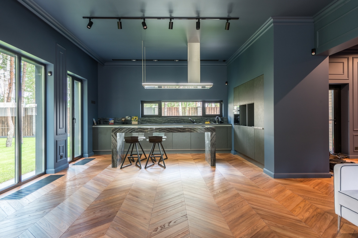 Luxury Home Flooring Ideas for Buckhead: Hardwood, Tile, and More 7 Modern kitchen with blue walls and ceiling, a marble island with two stools, luxury home flooring in wood herringbone, and large windows overlooking a green yard.