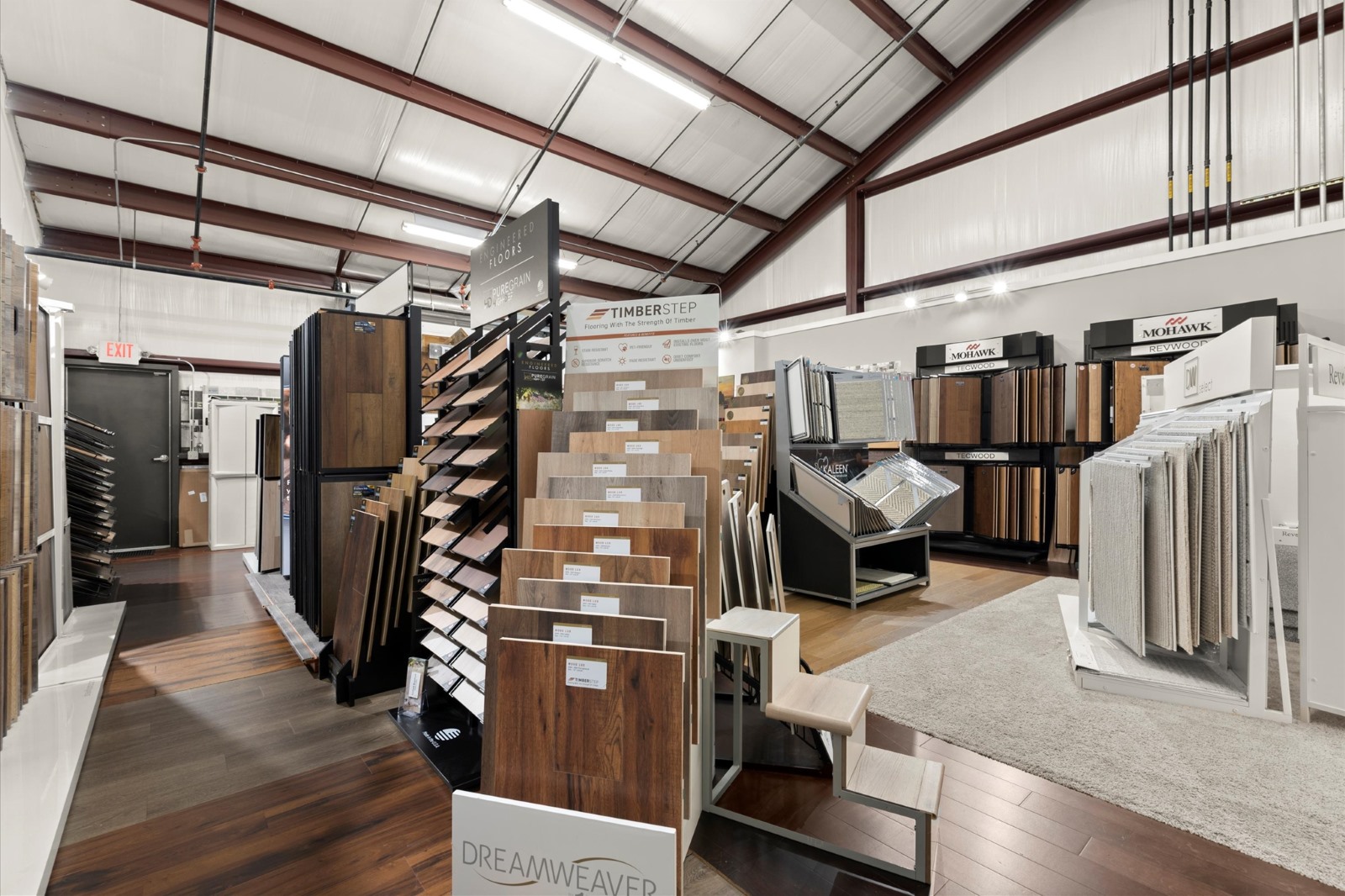 A flooring showroom displays various samples of hardwood, laminate, and carpet flooring on stands and racks under bright lighting, highlighting options available for different service areas.