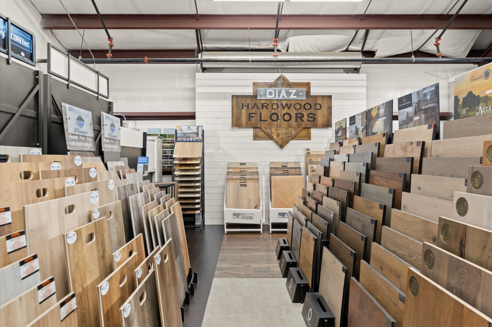 Hardwood Flooring 7 A showroom displays various hardwood flooring samples on racks, with a large “Diaz Hardwood Floors” sign mounted on the back wall.