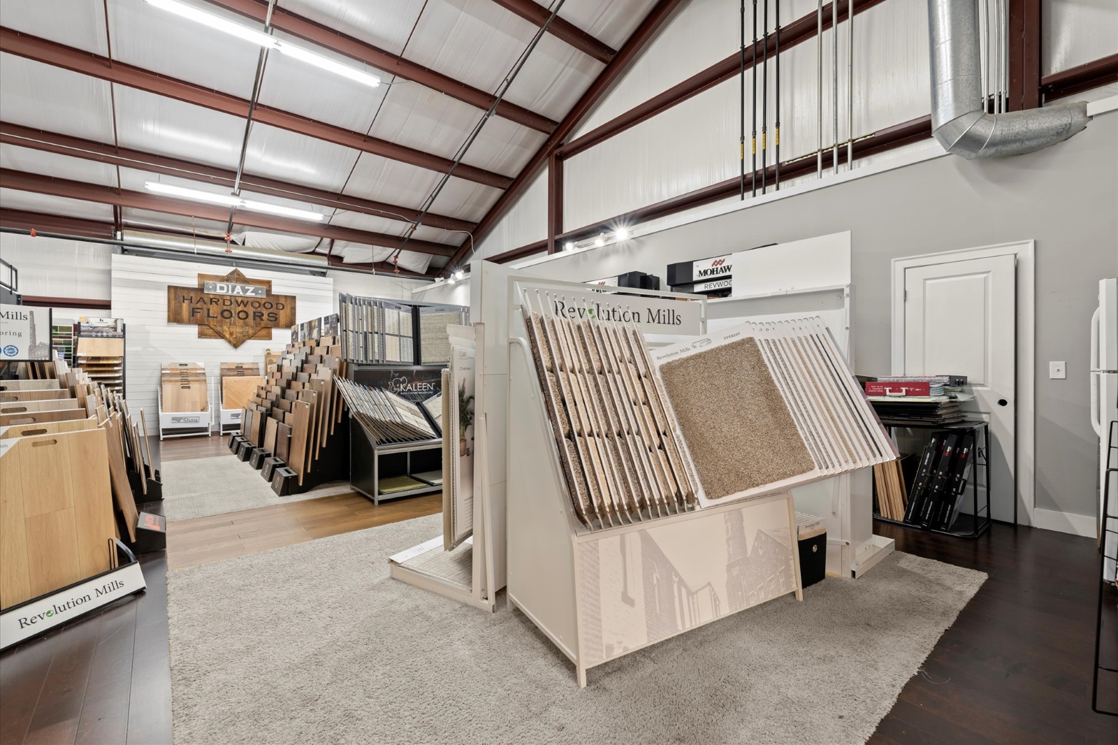 Showroom interior with carpet installation samples and hardwood floor options on display racks, metal ceiling beams, and a mix of flooring choices arranged neatly throughout the space.