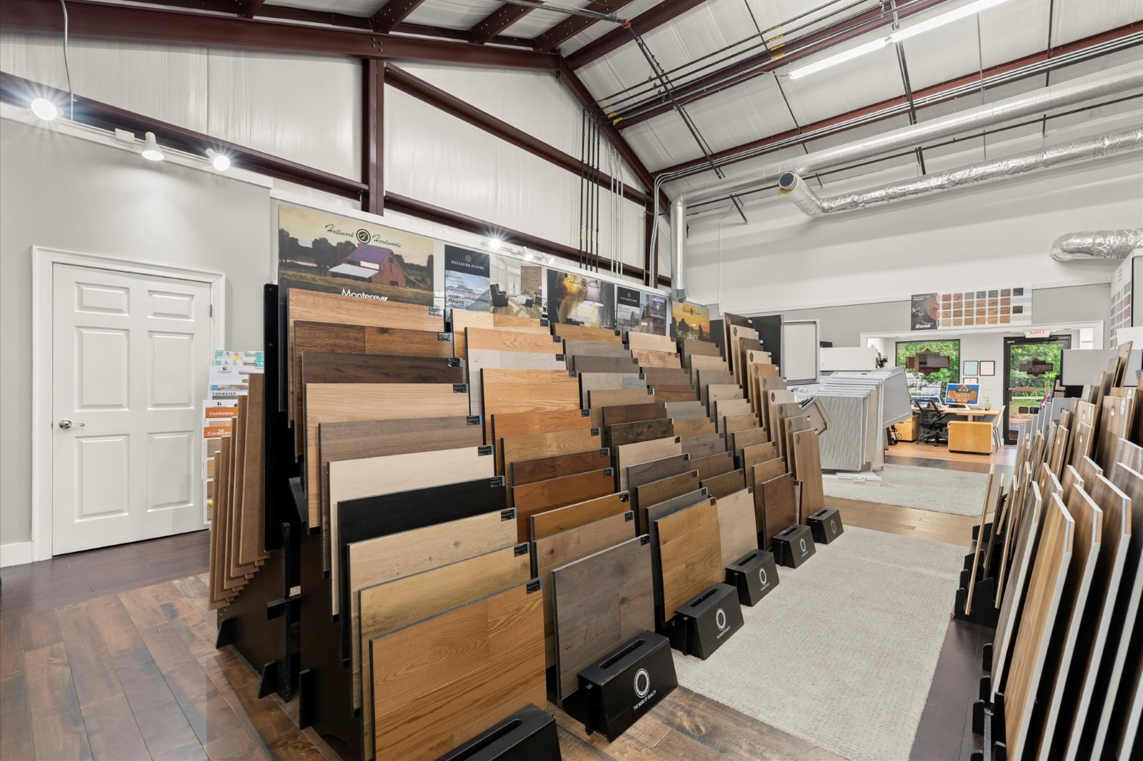 Showroom interior with multiple displays of wood and laminate flooring samples arranged in rows, under a high ceiling with exposed beams and ductwork.