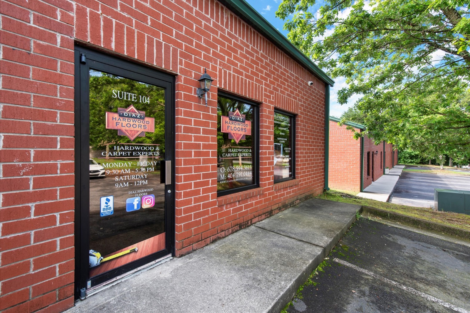 A brick commercial building with large windows and a glass door labeled "DIVAS HARDWOOD FLOORS." Business hours, contact details, and social media icons are displayed on the door. Trees and a parking lot are visible.