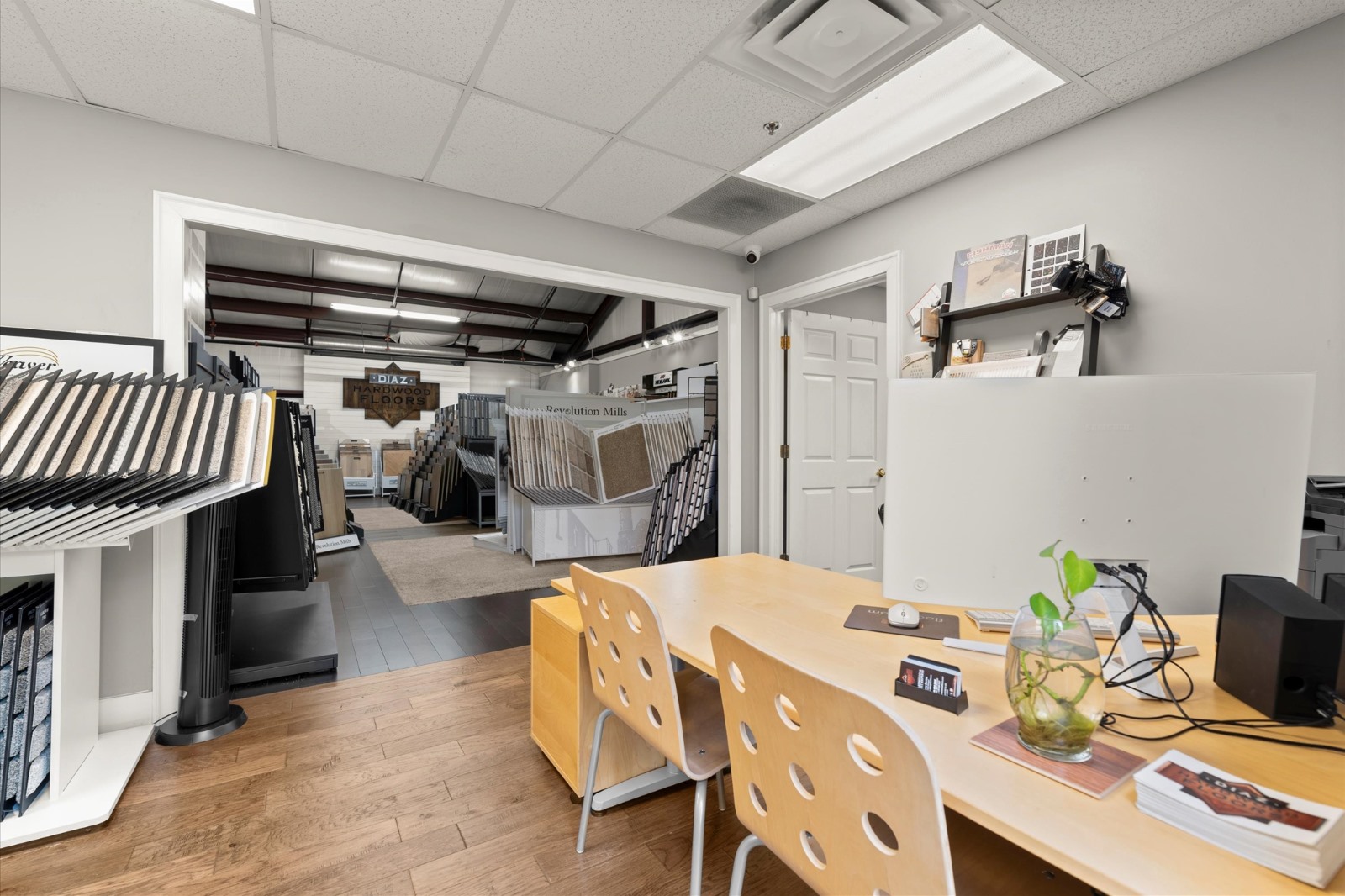 A showroom features display racks with hardwood flooring samples, a wooden desk with chairs, office supplies, and a plant in the foreground, all illuminated by fluorescent ceiling lights.