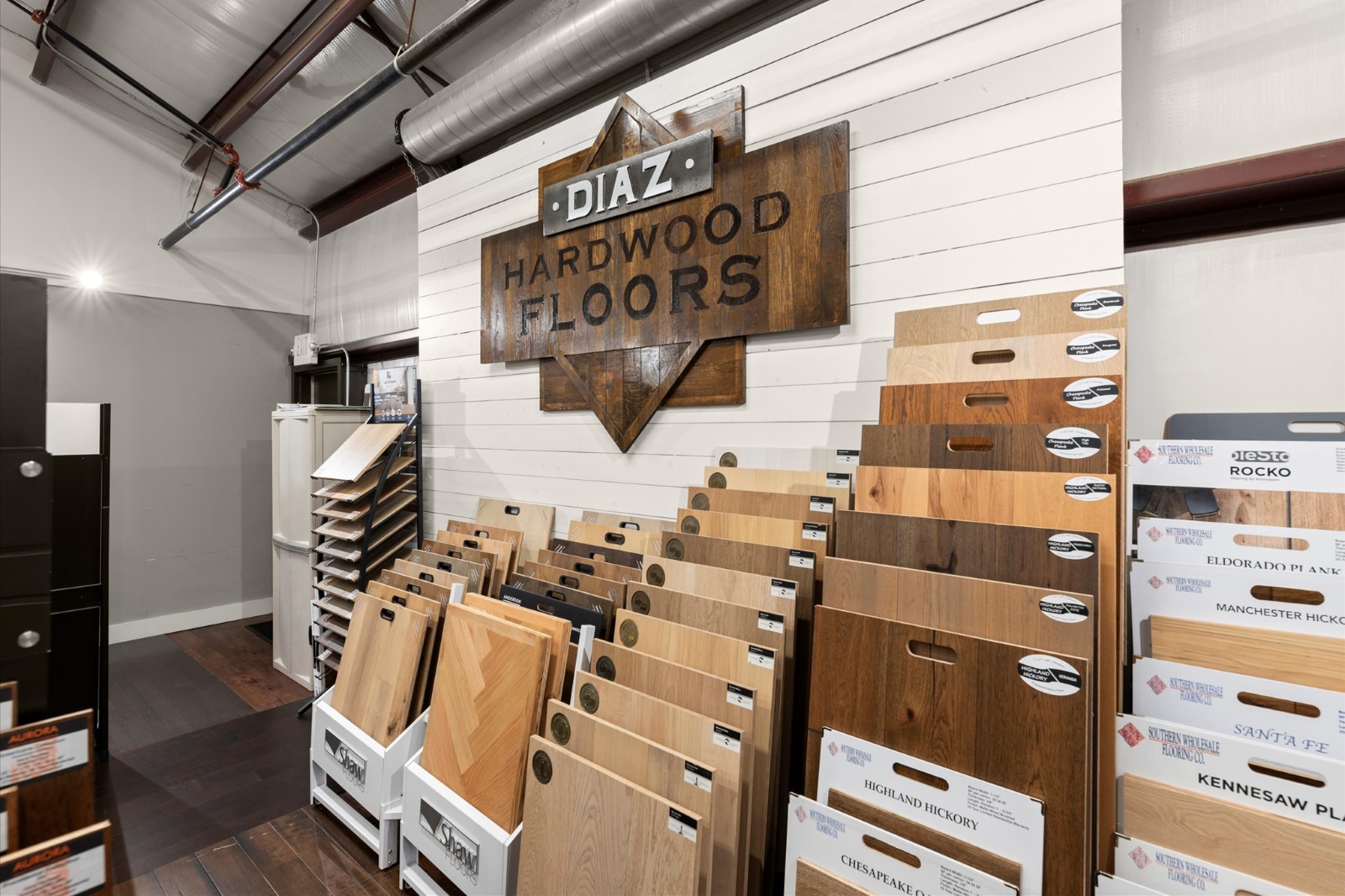 Display of various hardwood flooring samples arranged on stands beneath a large "Diaz Hardwood Floors" sign inside a showroom.