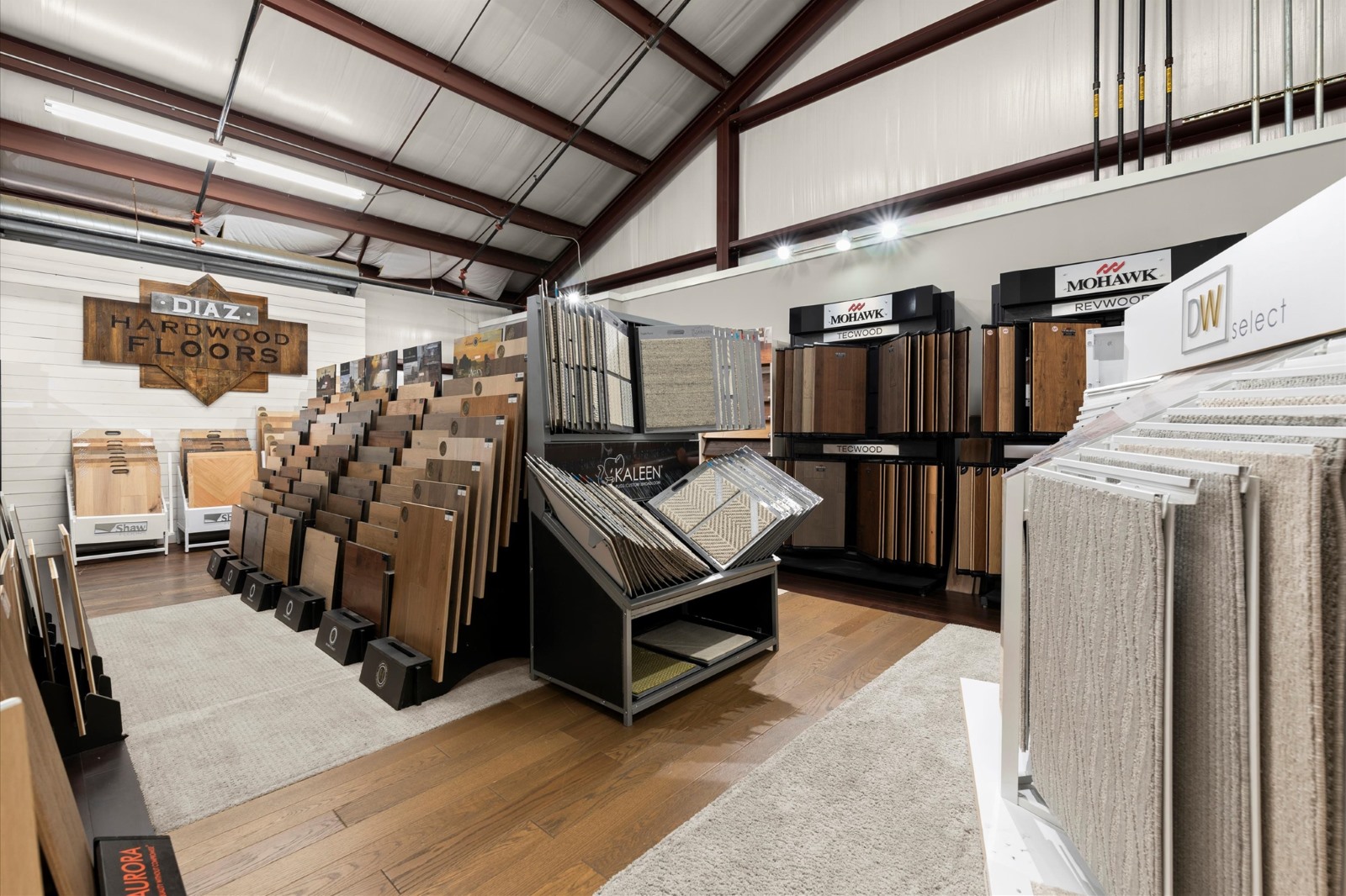 A showroom displays about various hardwood flooring samples, carpet samples, and wood floor panels arranged on racks and stands under a high metal ceiling.