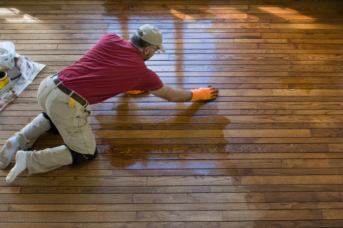 A man wearing gloves kneels on a wood floor, carefully applying a finish or stain, with part of the wood visibly darker and wet where he has worked.