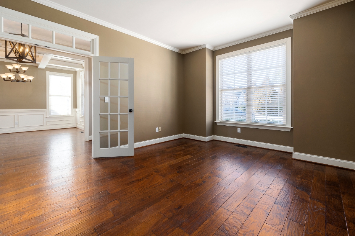Wood Floor Refinishing in Brookhaven: Key Signs It's Time to Refinish Your Floors 9 Empty room with wood floors, beige walls, white trim, a large window with blinds, and a glass-paned door leading to another room with a chandelier.