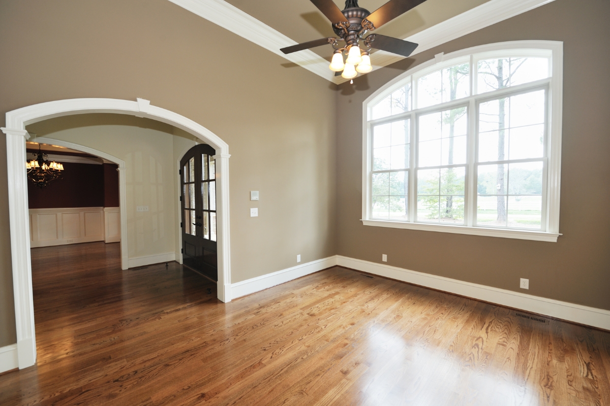 Wood Floor Finish Guide for Canton Homeowners 3 Empty room with wood floor finish, taupe walls, large arched window, ceiling fan, and an arched doorway leading to another room.