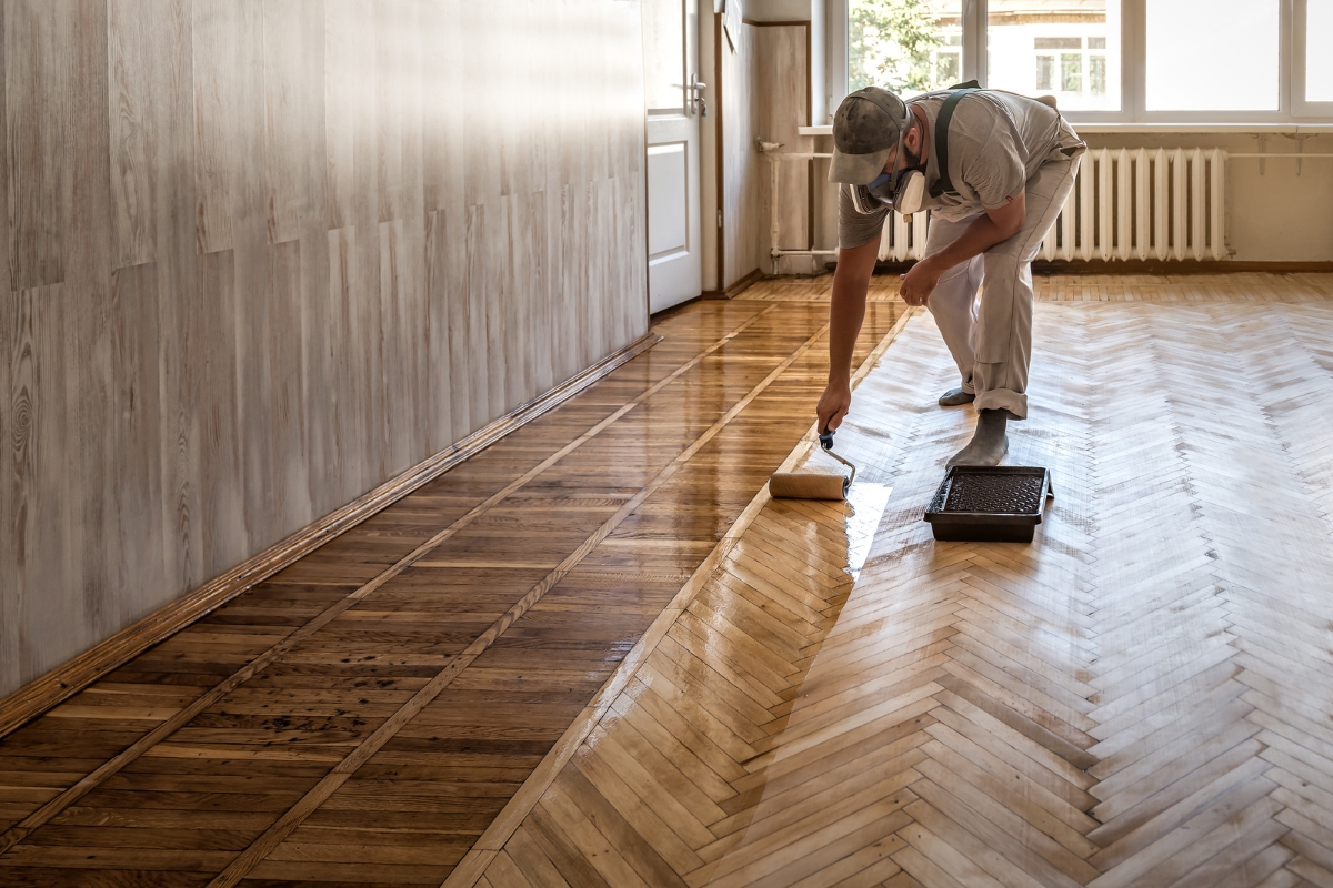 Wood Floor Finish Guide for Canton Homeowners 1 Person applying varnish to a wooden herringbone floor in a bright room, with half the floor already coated in a glossy wood floor finish.