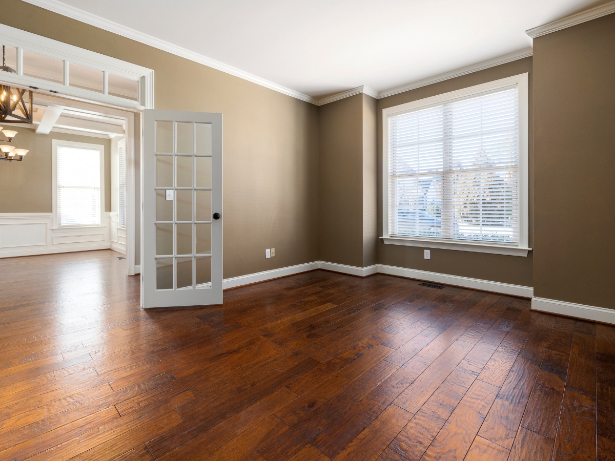 Engineered Hardwood vs. Solid Hardwood: Which is Better for Dallas Homes? 10 Empty room with brown walls, large window with blinds, engineered hardwood floor, and an open glass-paneled door leading to a bright adjoining space.