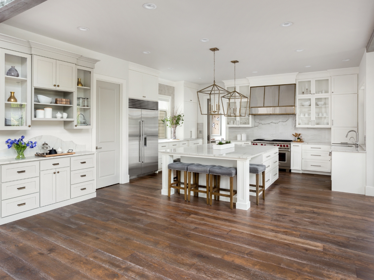 Engineered Hardwood vs. Solid Hardwood: Which is Better for Dallas Homes? 12 Spacious modern kitchen with white cabinets, stainless steel appliances, large island with stools, pendant lighting, and stylish engineered hardwood flooring.