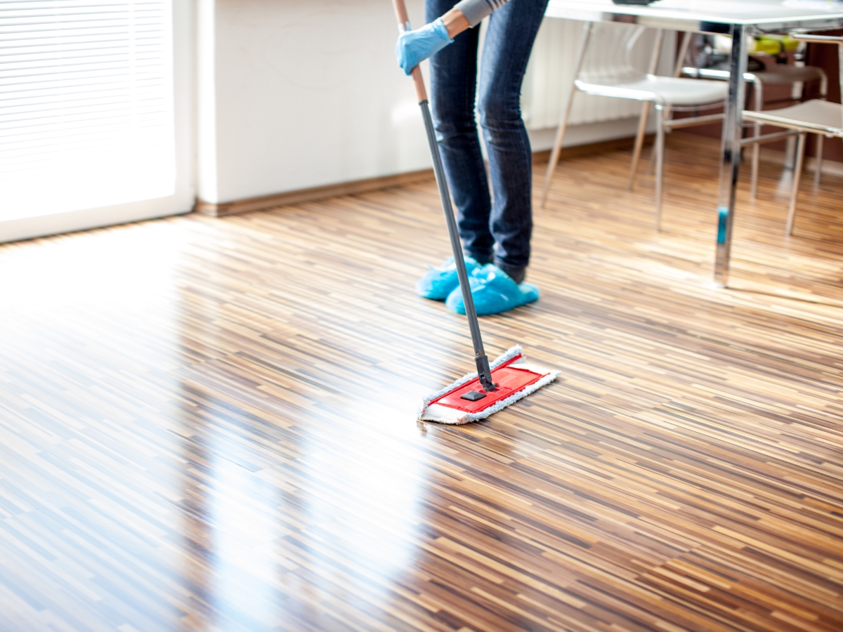 Engineered Hardwood vs. Solid Hardwood: Which is Better for Dallas Homes? 16 Wearing gloves and shoe covers, a person uses a flat mop to clean engineered hardwood flooring in a bright room with a table and chairs.