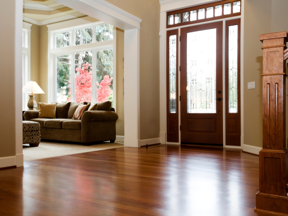 A spacious living room with engineered hardwood floors, a brown sofa by large windows, a lit table lamp, and a wooden front door with glass panels.