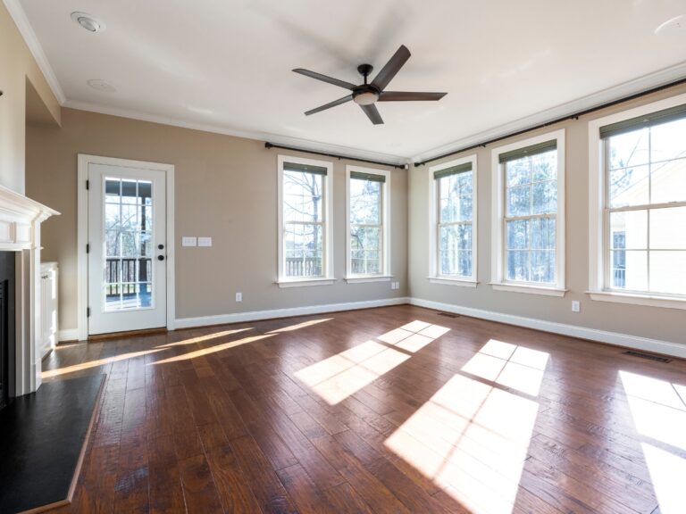 Bright, empty living room with restored hardwood floors, beige walls, multiple large windows, a ceiling fan, and a door leading to a porch.