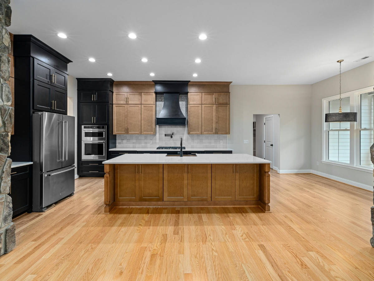 The Pros and Cons of Wide Plank Hardwood Flooring in Dunwoody GA Homes 3 Modern kitchen with wide plank hardwood floors, a central island, black and wood cabinetry, stainless steel appliances, and ceiling spotlights. Large window and pendant light at right.