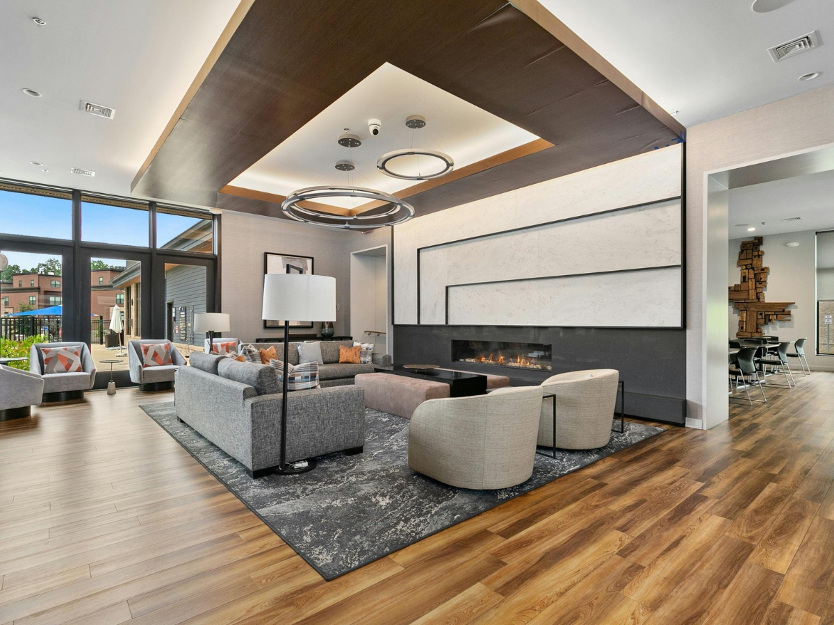 The Pros and Cons of Wide Plank Hardwood Flooring in Dunwoody GA Homes 4 Modern lounge area with gray sofas, armchairs, a large rug, wide plank hardwood floors, a linear fireplace, and floor-to-ceiling windows overlooking an outdoor area.