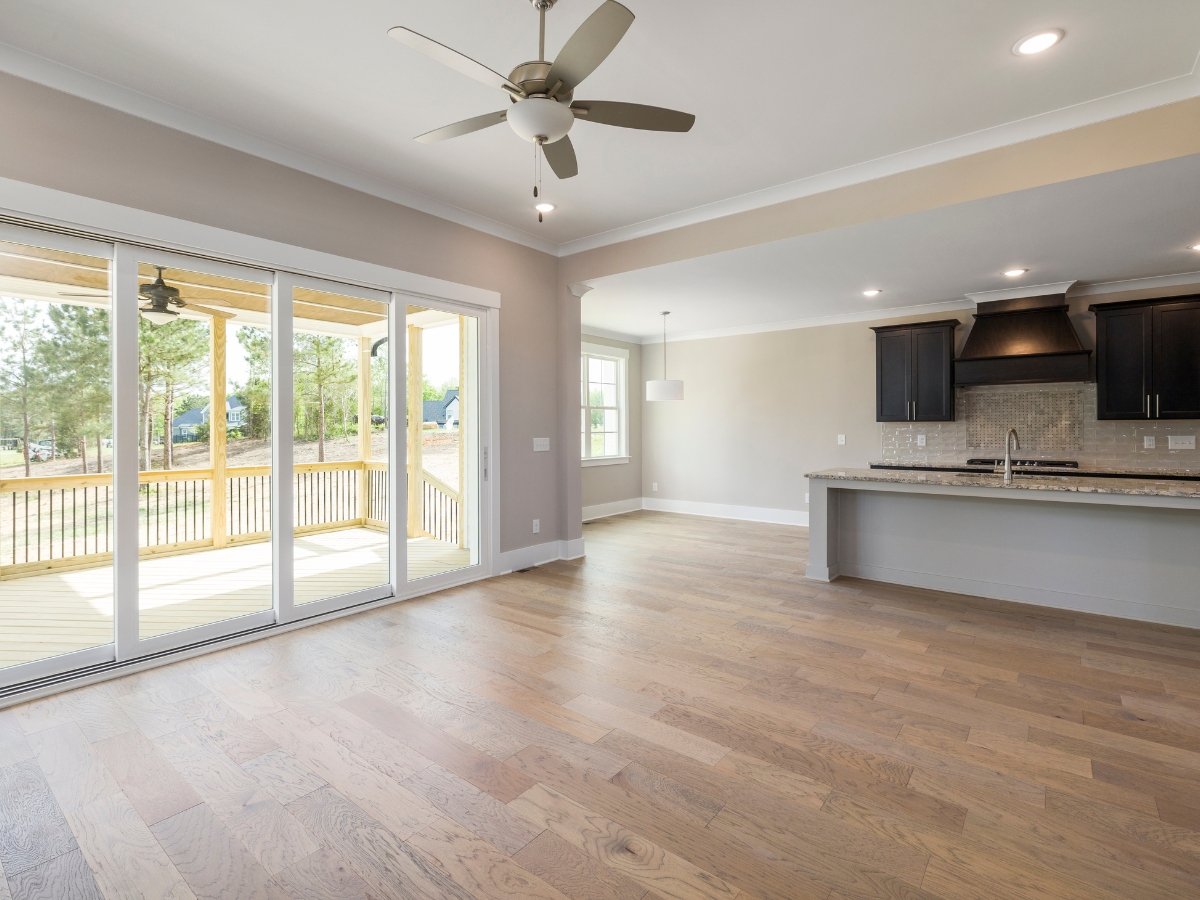 The Pros and Cons of Wide Plank Hardwood Flooring in Dunwoody GA Homes 1 Unfurnished open-plan living area with wide plank hardwood floors, ceiling fan, large sliding glass doors to a patio, and a modern kitchen with dark cabinets and a stone countertop.