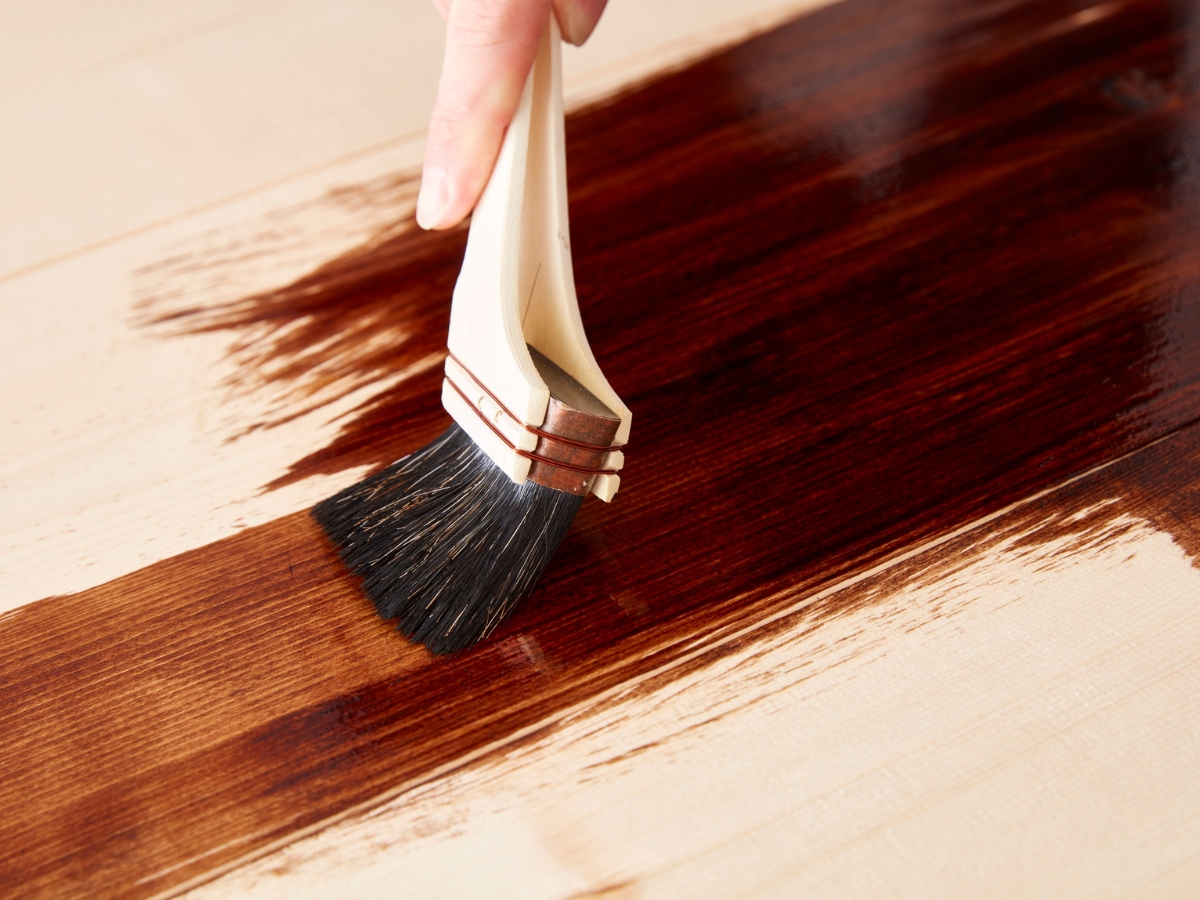 How to Choose the Best Stain Color for Hardwood Floors in Holly Springs 3 A hand uses a paintbrush to apply the best stain color for hardwood floors, coating a light-colored wooden surface with a rich, dark brown wood stain.