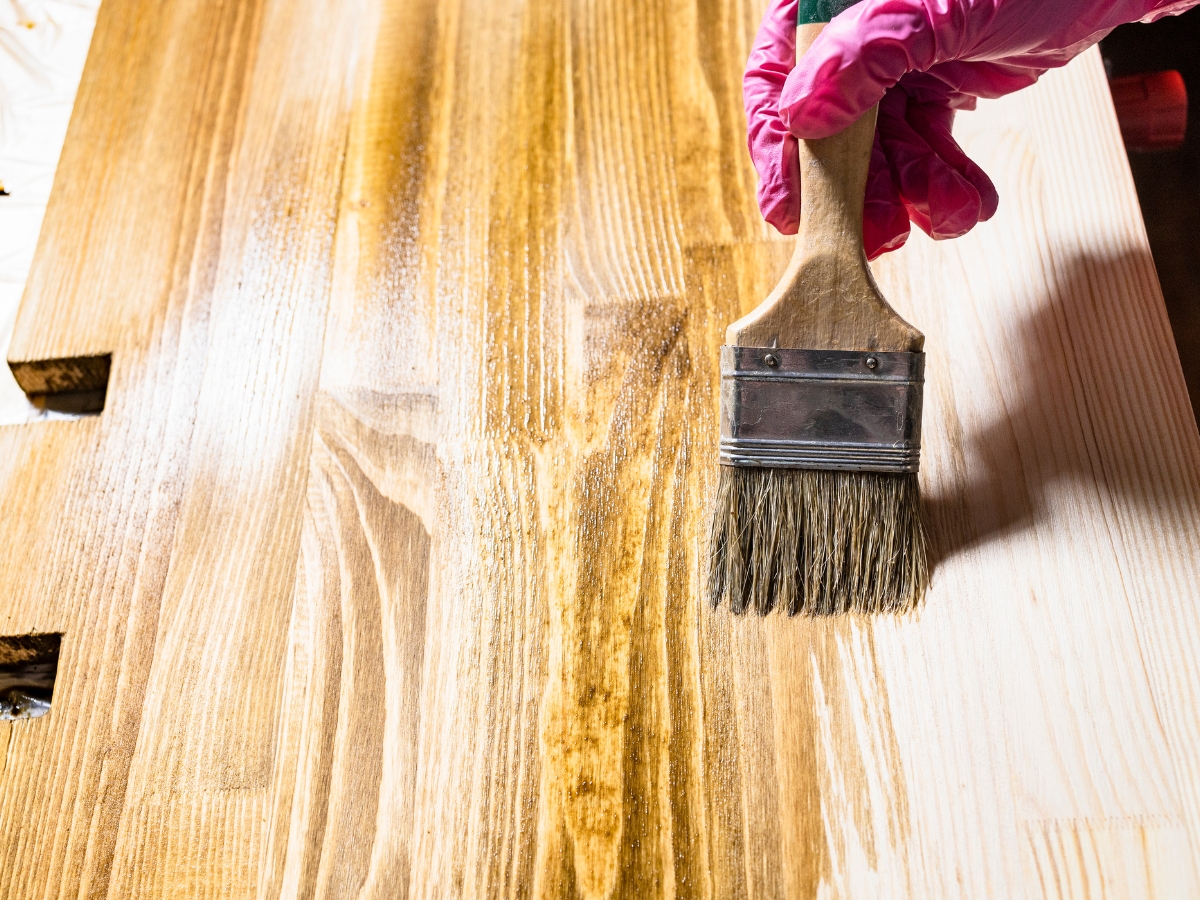 How to Choose the Best Stain Color for Hardwood Floors in Holly Springs 5 A hand in a pink glove uses a paintbrush to apply wood stain or varnish, demonstrating how to achieve the best stain color for hardwood floors on a wooden surface.