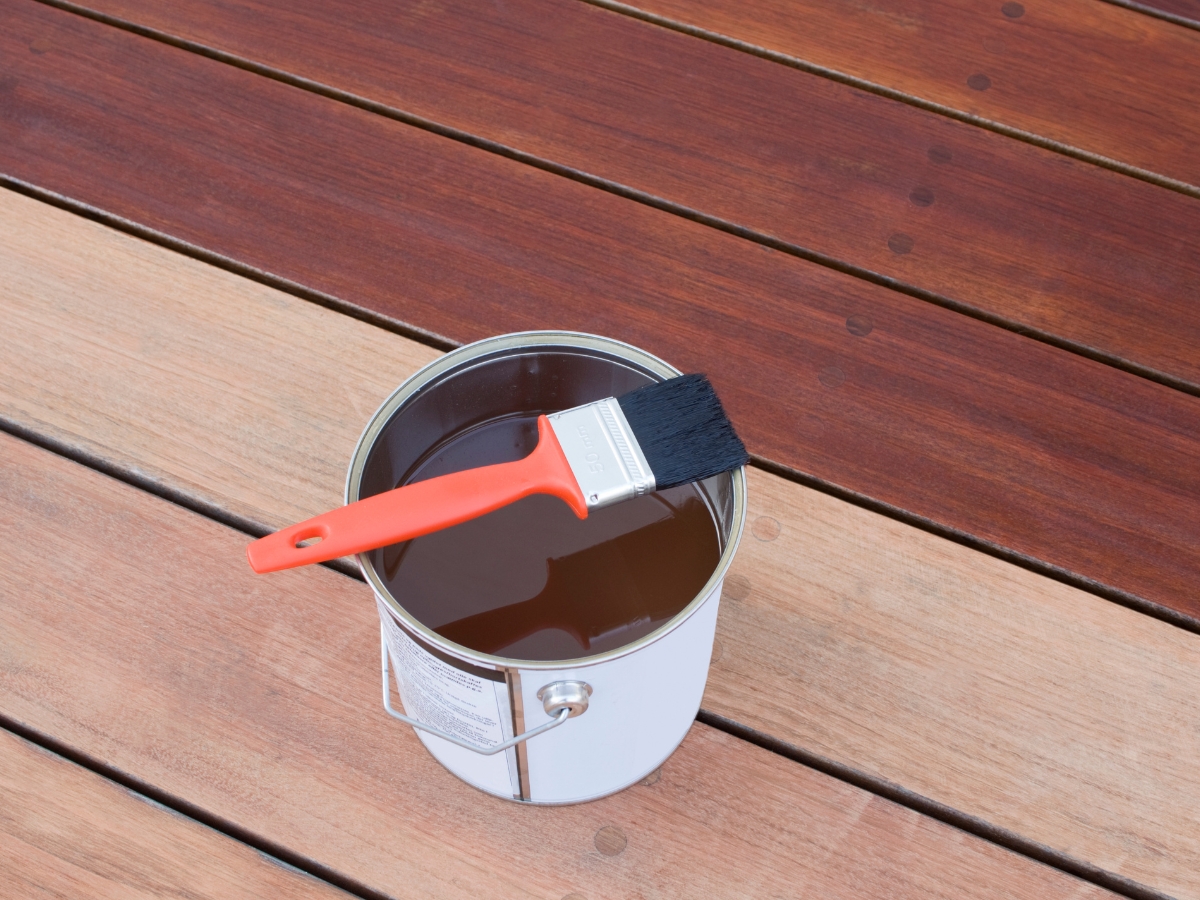 How to Choose the Best Stain Color for Hardwood Floors in Holly Springs 2 A paintbrush rests on top of an open can of wood stain, placed on a wooden deck with boards of varying shades, helping you explore the best stain color for hardwood floors.