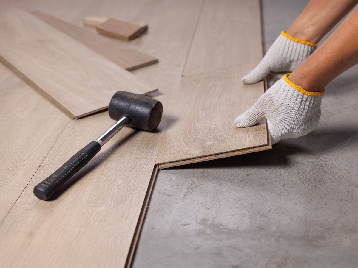 Johns Creek Home Flooring Guide: Hardwood, Laminate, or Vinyl? 2 Hands wearing gloves installing laminate flooring on a concrete surface with a rubber mallet and wood planks nearby, as featured in the Johns Creek Home Flooring Guide.