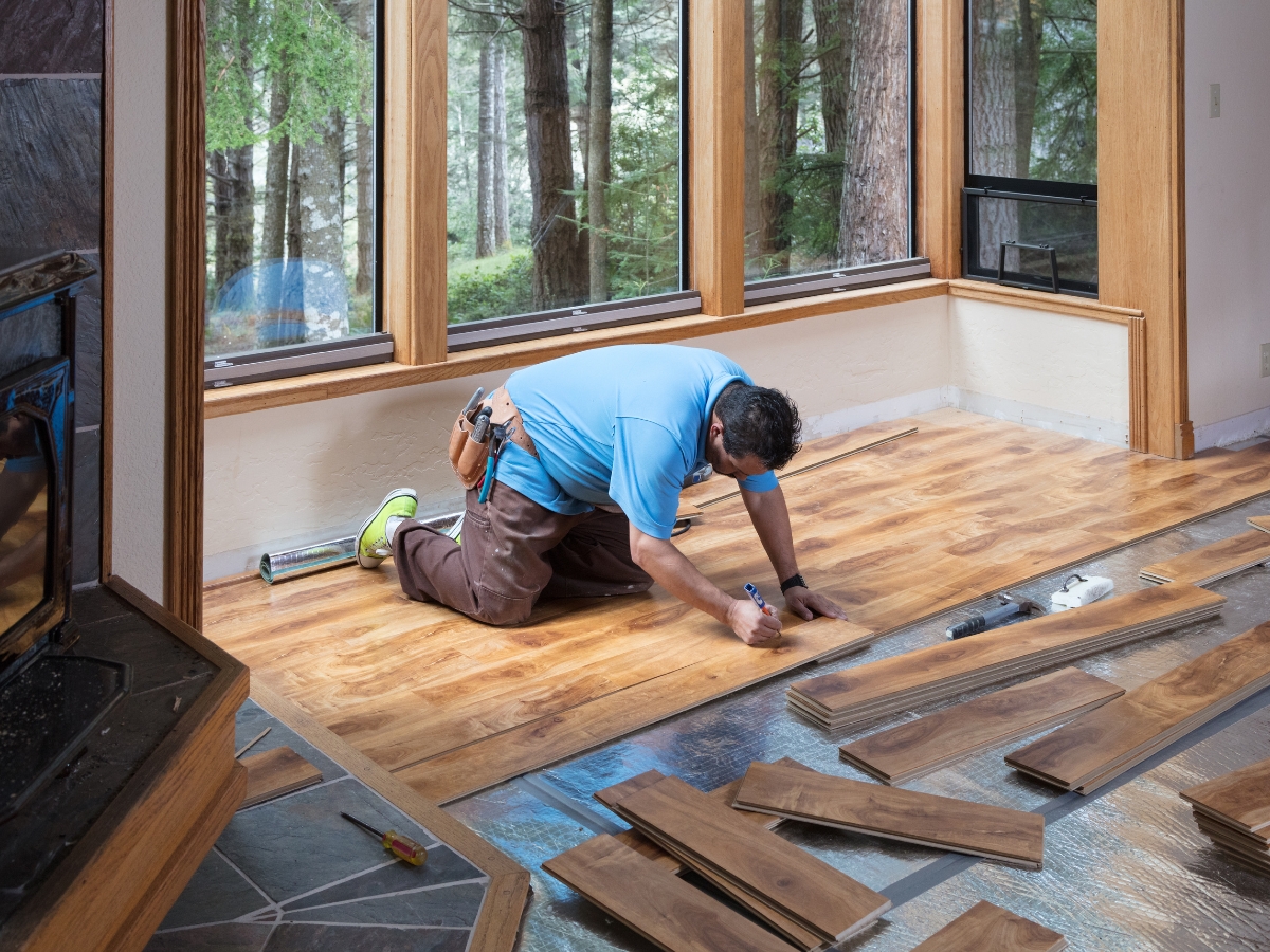Johns Creek Home Flooring Guide: Hardwood, Laminate, or Vinyl? 5 A man kneels on the floor measuring and installing wood flooring in a room with large windows overlooking trees, following tips from the Johns Creek Home Flooring Guide.
