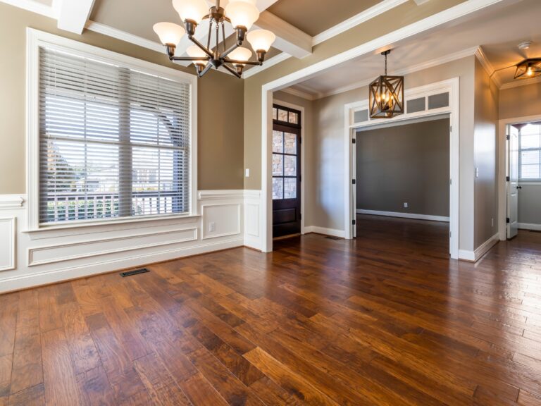 Empty room with large windows, wood flooring featured in our Johns Creek Home Flooring Guide, beige walls, white trim, and two ceiling light fixtures. Doorway leads to another room with similar features.