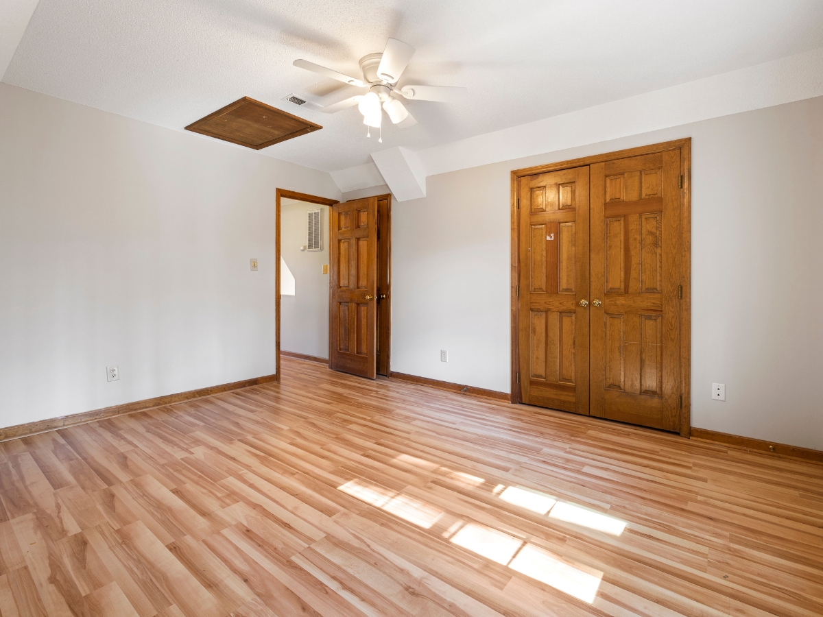 Empty room with light wood flooring—perfect for those looking to fix squeaky wood floors—beige walls, a ceiling fan, two wooden doors, and an attic access panel. Sunlight enters through a window outside the frame.