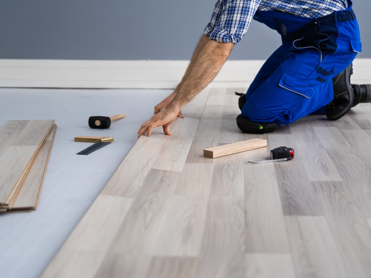 A person kneeling on the floor installs light wood laminate flooring, with tools such as a tape measure, mallet, and ruler nearby—essential steps to fix squeaky wood floors and ensure a smooth, quiet finish.