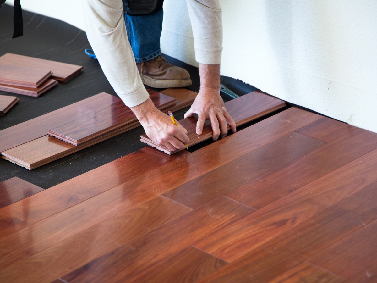 A person measures and installs wooden flooring planks, kneeling on the floor with tools and uninstalled planks nearby—an essential step to fix squeaky wood floors.