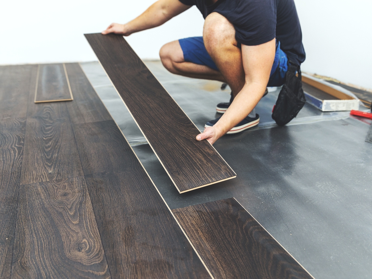 A person wearing blue shorts installs dark wood laminate flooring—one of the best flooring upgrades before selling in Mableton, GA—carefully positioning a plank into place on a partially finished floor.