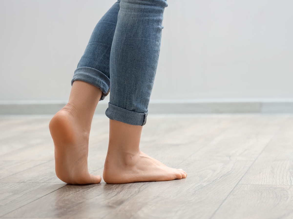 Person standing barefoot on a wooden floor with one foot flat and the other raised onto the toes, wearing rolled-up jeans—demonstrating mindful laminate care by keeping the surface clean and scratch-free.