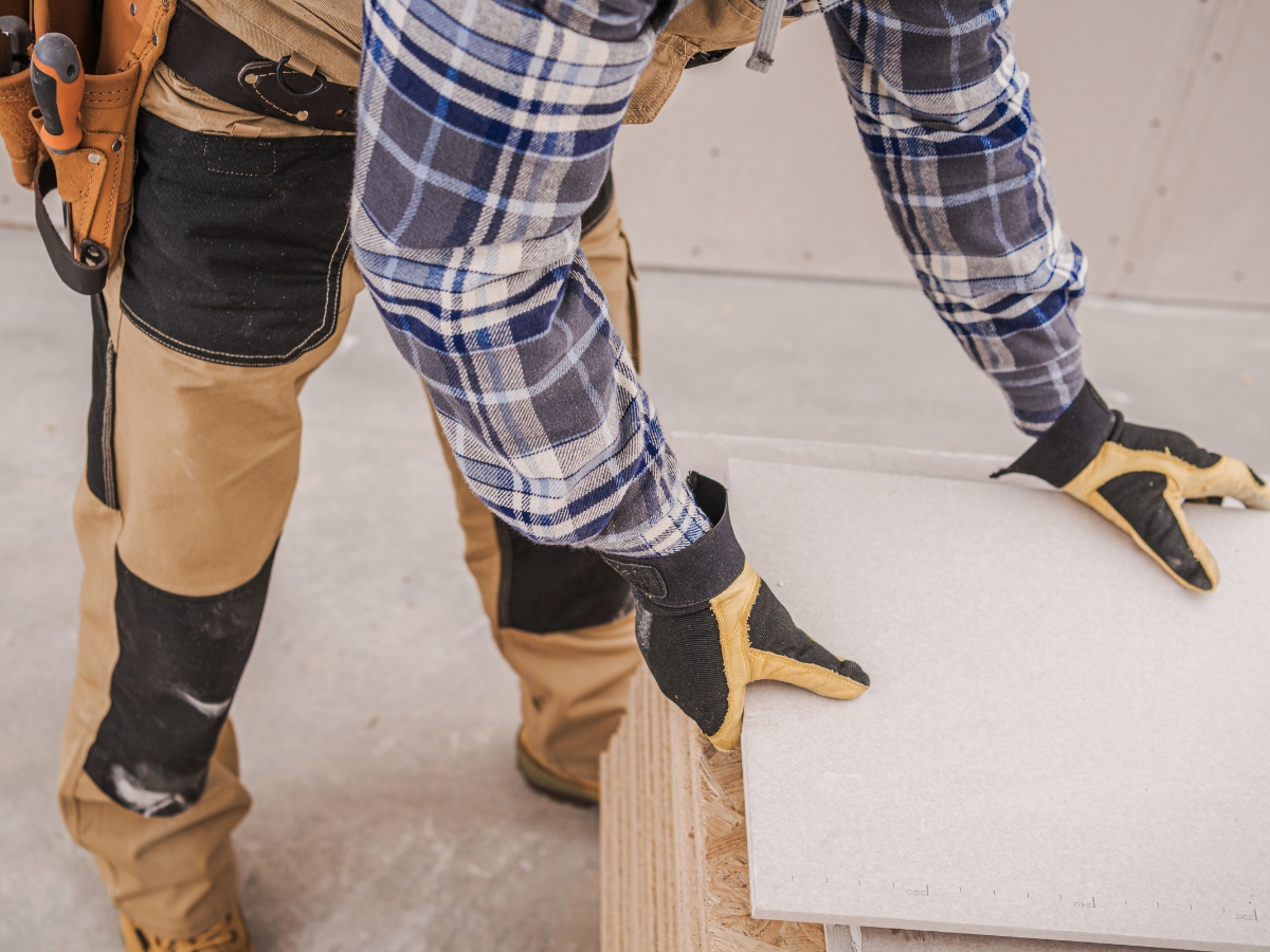 Person wearing gloves and a plaid shirt preparing a sheet of drywall on a wooden surface at a construction site, ideal for finishing small spaces efficiently.
