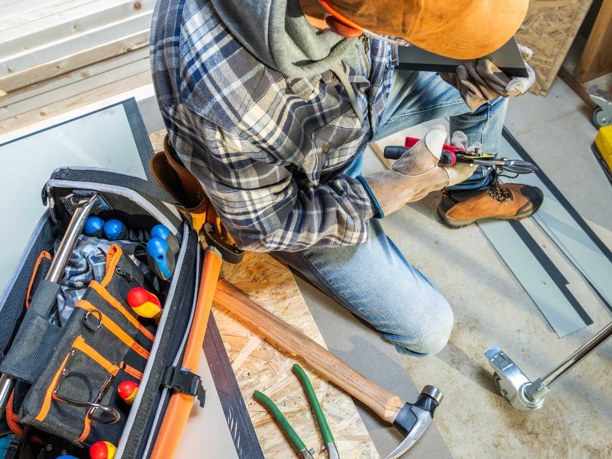 Person in workwear using pliers on a metal strip next to a toolbox, hammer, and various hand tools on a wooden surface in a workshop designed for working efficiently in small spaces.