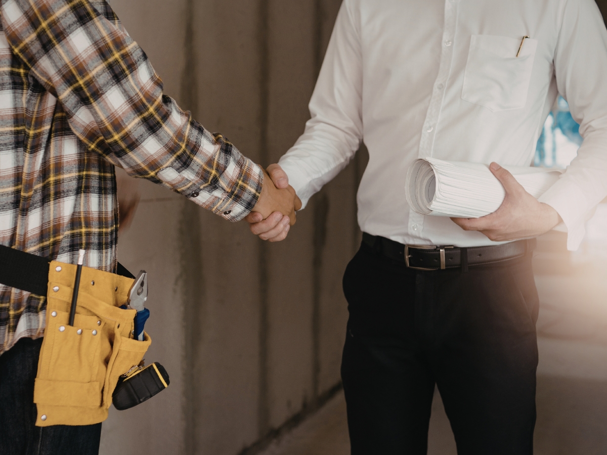 How Transitional Flooring Blends Traditional and Modern Style 4 A person in a plaid shirt with a tool belt shakes hands with another person in a white shirt holding rolled construction plans, likely discussing transitional flooring options for their latest project.