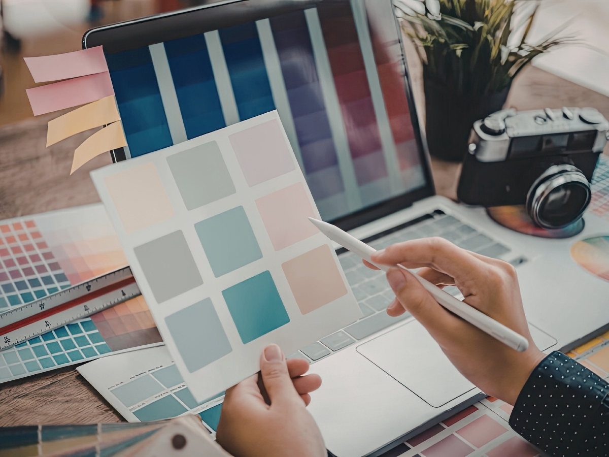 A person holds a color swatch card and pen in front of a laptop displaying a similar palette, with a camera, ruler, and color charts nearby for precise color matching.