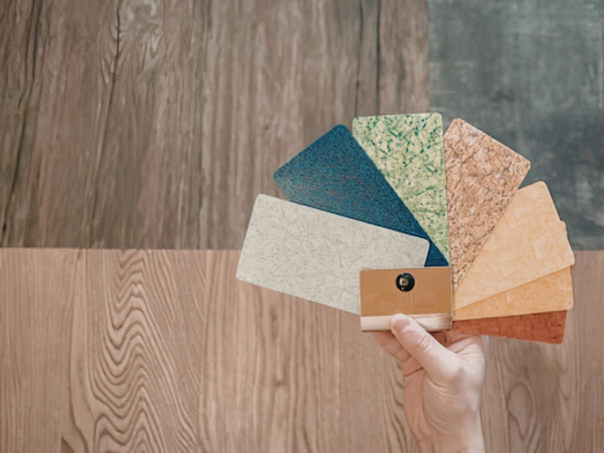 A hand holds a fan of flooring material samples in various colors and textures above a wood-patterned floor, showcasing options for perfect color matching.