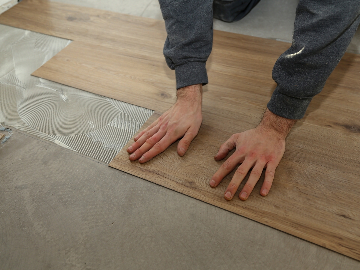 Person installing wood-look vinyl flooring planks over adhesive on a cement floor, carefully color matching each plank before pressing it firmly into place with both hands.