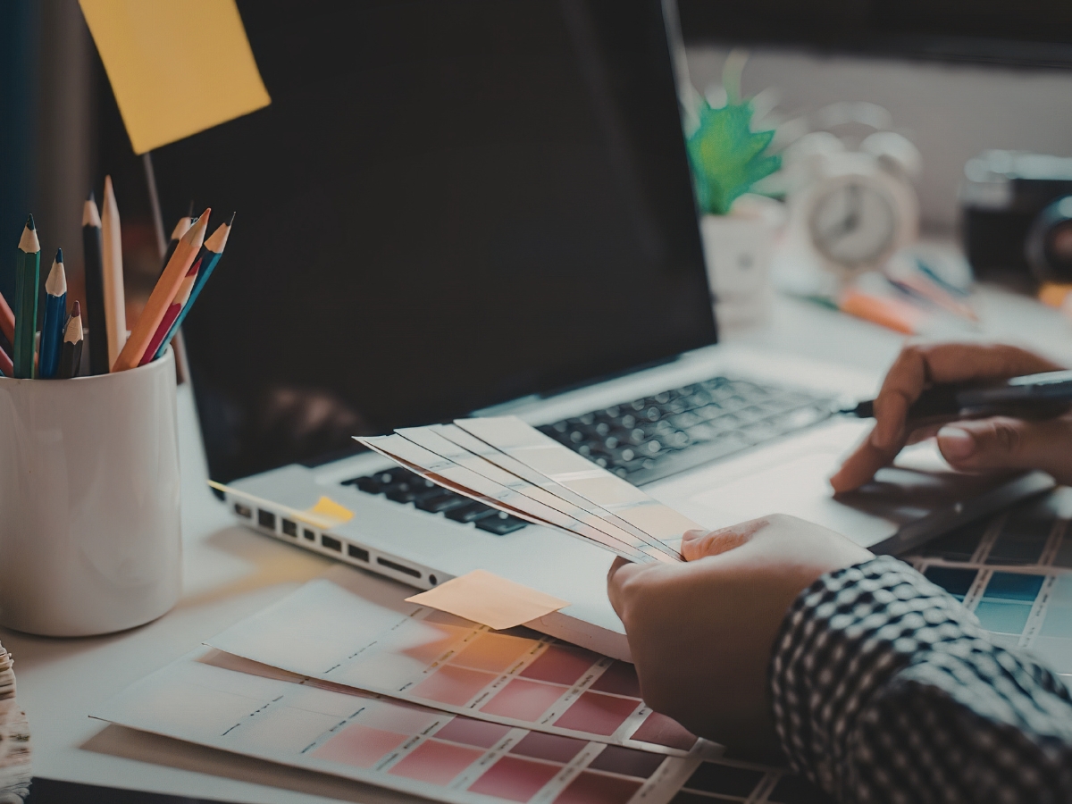 Person reviewing color swatches at a desk, focusing on color matching with a laptop, a cup of colored pencils, sticky notes, and other office supplies visible.