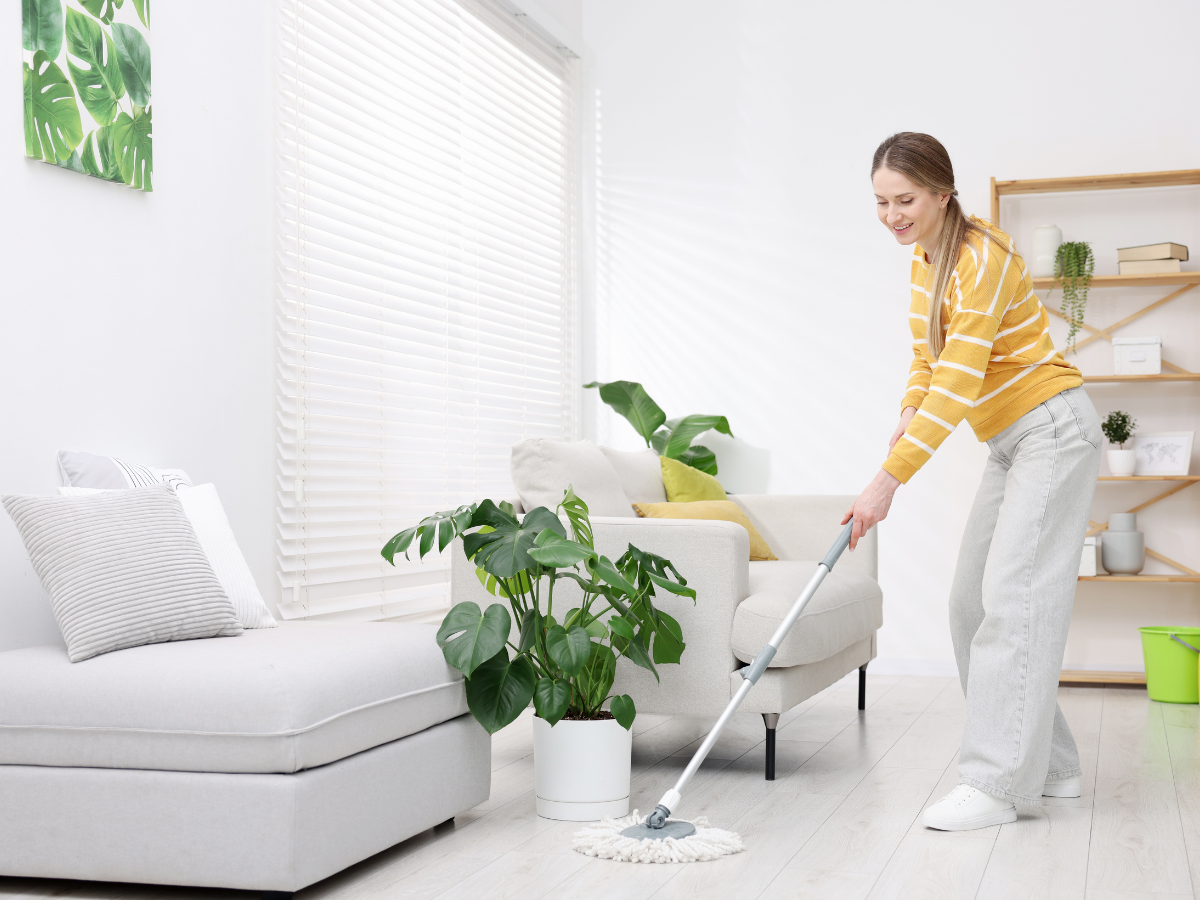 A woman in a yellow striped sweater mops the hypoallergenic flooring in a bright, modern living room with plants and neutral-colored furniture.