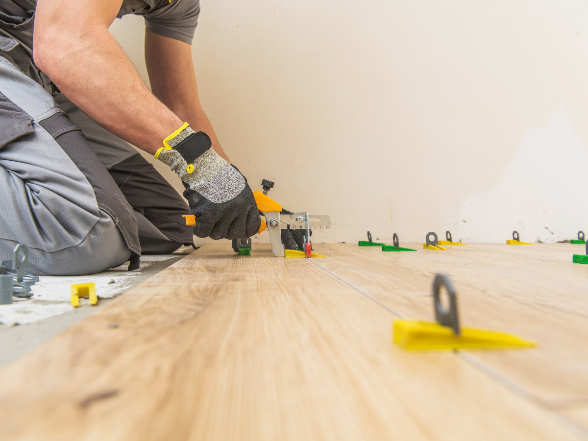 Person installing wood-look hypoallergenic flooring tiles using spacers and a leveling tool. The worker is wearing gloves and kneeling on the floor.