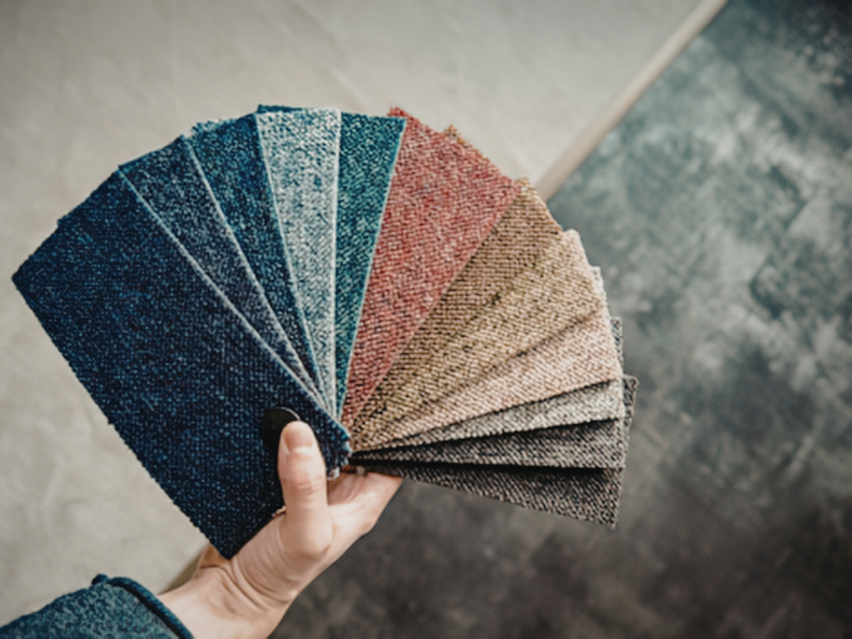 A hand holds a fan of textured fabric swatches in various shades of blue, red, brown, and gray against a neutral background—ideal for enhancing both décor and acoustics.